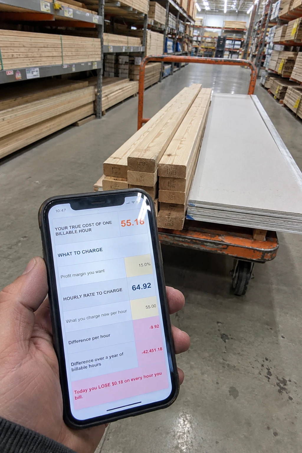 A contractor's hand holding a phone with the True Price Calculator in the lumber aisle of a hardware store